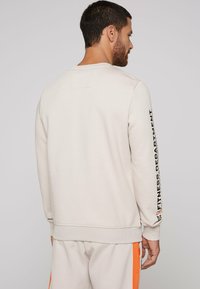 Beige bomulds sweatshirt med lange ærmer, ribbet kant og manchetter samt trykt tekst langs den højre ærme. Glat tekstur.