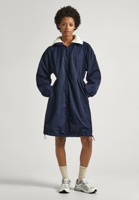 Manteau bleu marine, de longueur genou, avec un col en fausse fourrure blanche. Il présente un zip à l'avant, des poignets élastiques et un ourlet à cordon pour un ajustement personnalisable.