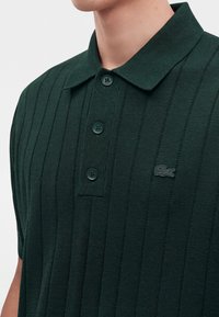 Polo en maille côtelée vert foncé avec trois boutons et logo crocodile sur la poitrine porté par une personne vue du cou vers le bas.