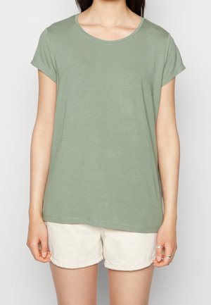 Frau trägt ein hellgrünes T-Shirt mit kurzen Ärmeln und weiße Shorts, steht mit entspannten Armen an den Seiten vor einem einfachen Hintergrund.