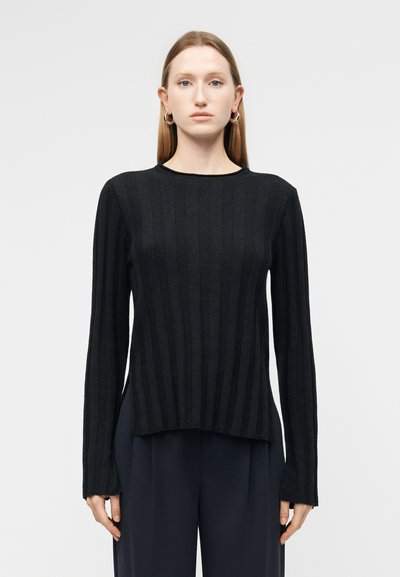 Femme aux cheveux lisses et clairs portant des boucles d'oreilles créoles en or, un pull noir côtelé à manches longues et un pantalon large et sombre, debout devant un fond uni.