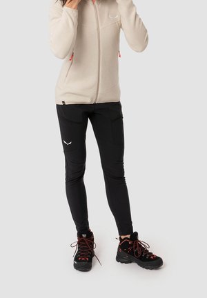 Person wearing beige zip-up fleece, black fitted outdoor pants, and black hiking boots with red laces, standing against plain background.