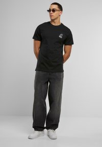 Svart bomull T-skjorte med et lite hvitt grafisk design på brystet, kombinert med mørkegrå denimbukser og hvite joggesko. Modellen står og poser.