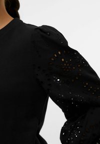 Svart sweatshirt med puffärmar. Den strukturerade tyget har utskurna blommönster. Den släta kroppen kontrasterar med den detaljerade ärmdesignen.