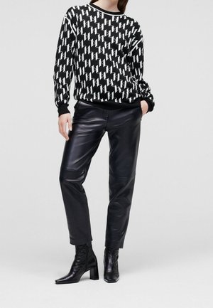Kobieta ubrana w czarno-biały sweter w wzory, czarne skórzane spodnie i czarne botki na obcasie, pozująca na jednolitym tle.