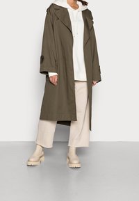 Abrigo trench verde oliva con solapas anchas, corte holgado y mangas abotonadas. Combinado con una sudadera blanca con capucha, pantalones beige de pierna ancha y botas gruesas beige.