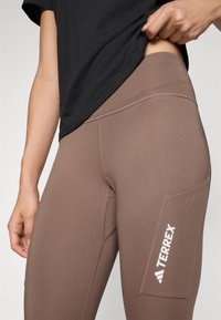 Brune leggings med en jevn tekstur, høy midje og sidelomme, med en hvit "TERREX"-logo og avrundet design.
