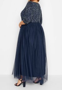 Tyrkysově modré večerní šaty se zdobenou korzetovou částí, tříčtvrtečními rukávy a splývavou tylovou sukní. Obsahují také sandály na kotníkovém pásku.