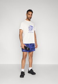 Λευκό μπλουζάκι με μπλε γραφικό βουνού, συνδυασμένο με μπλε σορτς με σχέδια. Μαύρα αθλητικά παπούτσια και φιάλη νερού ολοκληρώνουν την αθλητική εμφάνιση.