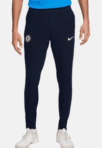 Pantaloni sportivi blu navy in tessuto a coste, con logo del Chelsea FC e swoosh bianco Nike. Design aderente con vita elastica.
