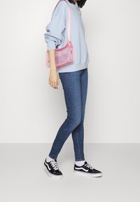 Persona en sudadera azul y jeans ajustados, llevando zapatillas negras y blancas, y una bolsa de hombro transparente rosa.