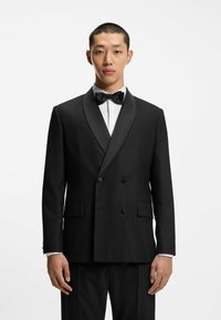 Giacca da smoking nera doppiopetto con rever in raso, abbinata a una camicia bianca e un papillon nero, dotata di tasche laterali.