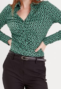 Femme portant une chemise à motif vert à boutons, rentrée dans un pantalon noir avec une fine ceinture noire, les mains sur les hanches.