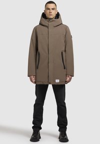Bruine geïsoleerde parka met hoge kraag, ritszakken en capuchon. Heeft zwarte accenten en een logopatch aan de onderrand.
