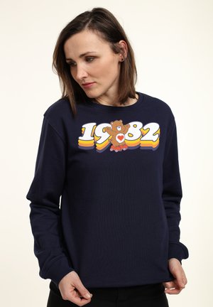 Marineblå sweatshirt lavet af bomuld, med et multifarvet grafisk design med en bjørn og året "1982" trykt på forsiden. Lange ærmer.