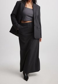 Frau, die ein dunkel gestreiftes Blazer und einen passenden langen Rock trägt, kombiniert mit einem grauen Cropped-Top und schwarzen, spitzen Slippern.