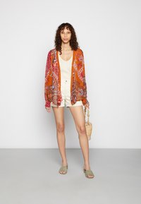 Jaqueta estilo quimono em laranja com padrões paisley, combinada com uma blusa bege e calções brancos. Segurando uma bolsa de palha, vestindo sandálias de slide verdes.