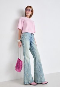 Lichtroze geribbelde top met korte mouwen, vervaagde blauwe flared jeans met bloemenversieringen, roze stiletto's en een roze franje handtas.