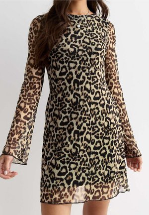 Femme portant une robe courte à manches longues imprimée léopard avec un tissu transparent et des colliers argentés superposés, sur un fond uni.