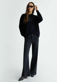 Felpa oversize blu navy con logo in rilievo, abbinata a jeans neri a vita alta e a campana e a ballerine nere a punta con texture. Occhiali da sole neri.