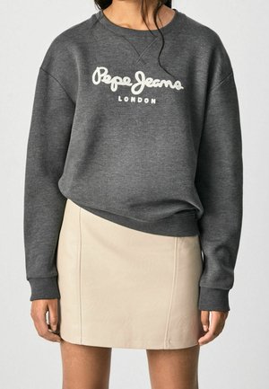 Kobieta ubrana w ciemnoszarą bluzę Pepe Jeans London i beżową skórzaną mini spódnicę, stojąca na jednolitym tle.