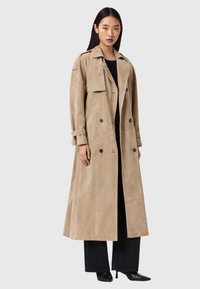 Μπεζ παλτό τύπου τρανς από σουέντ με διπλό κούμπωμα, τσέπη με καπάκι και μανίκια γυρισμένα, σε συνδυασμό με μαύρο παντελόνι φαρδύ και ψηλοτάκουνα παπούτσια.