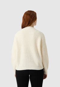 Kremowy sweter dziany o luźnym kroju, z prążkowaną fakturą i opadającymi szwami ramion, noszony na ciemnych legginsach.