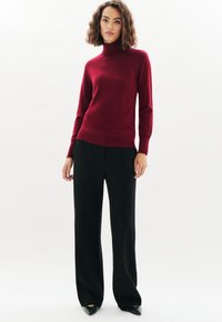 Vrouw met schouderlang, krullend haar die een bordeauxrode coltrui, zwarte wijde broek en zwarte puntige schoenen draagt.