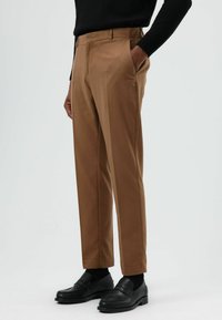 Bruine broek met een getailleerde pasvorm, voorzien van zijzakken en een gladde textuur; gecombineerd met zwarte loafers en sokken.