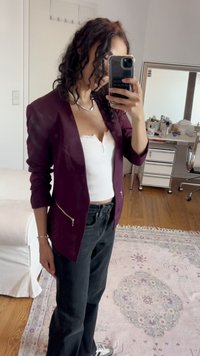 Bordeaux blazer met zilveren ritsaccenten, gedragen over een witte top en gecombineerd met zwarte jeans. De stof lijkt glad en heeft een aansluitend ontwerp.