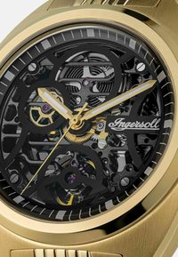 Goudkleurig mechanisch horloge met een zwart skelet-dial, dat intrikate tandwielen en paarse accenten toont, en voorzien is van zilveren uurmarkeringen.