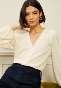 Blusa bianca con maniche a sbuffo, pois testurizzati e bordo in pizzo sul collo. Abbinata a jeans di jeans scuri.