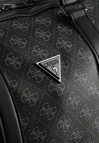 Bolso negro texturizado con patrón de monograma gris claro repetido y placa triangular con el logo de Guess en el panel frontal cerca de la hebilla de la correa.