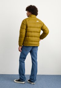 Gulgrønne oppustelige jakke med høj krave, quiltet design og The North Face-logo på ryggen, parret med blå jeans og sneakers.