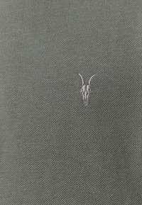 Khaki-grüner Stoff mit einem strukturierten Strickmuster, das ein zentral platziertes hellgraues gesticktes Logo eines gehörnten Tieres zeigt.