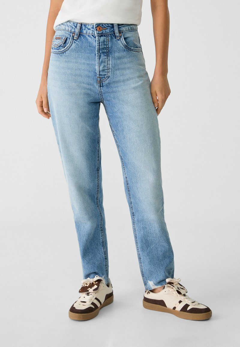 Jean droit en denim bleu clair avec une taille moyenne, cinq poches et un léger délavage. Assorti à des baskets beiges et brunes.