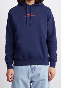 Mörkblå hoodie i mjukt material, med en framficka, dragsko på huvan och tydlig orange branding på bröstet.