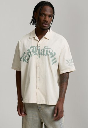 Joven con trenzas, vestido con una camisa beige de manga corta abotonada y vaqueros de lavado claro sobre un fondo gris liso.