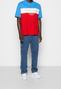 T-shirt a maniche corte con parte superiore blu, parte inferiore rossa, striscia bianca e logo dorato "Tommy Jeans". Indossato con jeans di denim blu e sneaker bianche.