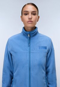 Giacca in pile blu chiaro con colletto alto, zip frontale completa e patch logo tono su tono sul petto. Presenta una superficie testurizzata e tasche laterali.