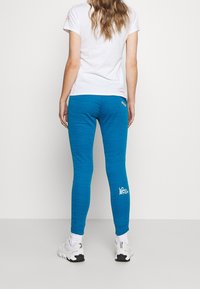 Blue active leggings with a textured pattern and white logo detail; paired with a white T-shirt and athletic shoes, viewed from the back.