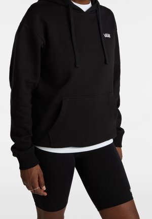Person trägt einen schwarzen Vans-Hoodie mit Fronttasche und Kordelzug, kombiniert mit schwarzen Vans-Radshorts und einem weißen Unterhemd.