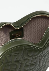 Borsa in pelle verde con motivi geometrici in rilievo, cerniera argentata parzialmente aperta, che rivela un interno in tessuto stampato e un piccolo etichetta in pelle.