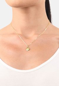 Primo piano di una collana d'oro con un piccolo pendente rotondo indossata al collo di una persona sopra una maglietta bianca.