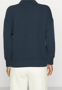 Felpa blu navy, tessuto morbido, vestibilità comoda, collo alto, spalle scivolate, polsini e orlo a costine. Indossata con pantaloni chiari.