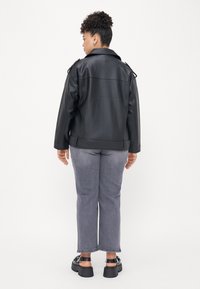 Black faux leather biker jacket with shoulder epaulettes, standing collar, and side pockets, paired with gray denim trousers and chunky black sandals.