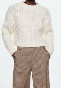 Dikke crème gebreide trui met een geribde ronde hals en kabelpatronen. Gecombineerd met hooggeplaatste geblokte broek met knoopdetails.