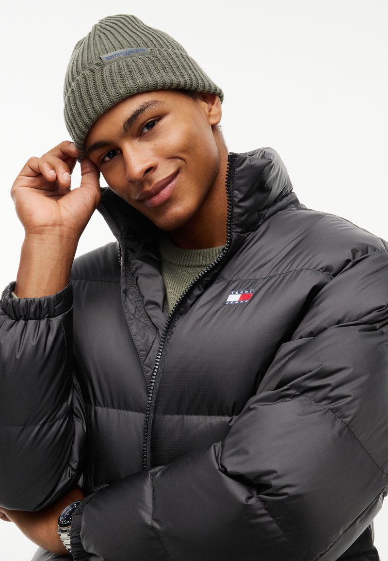 Jeune homme portant un bonnet en maille gris Tommy Jeans et une doudoune noire avec le col relevé, souriant et touchant le bord du bonnet.