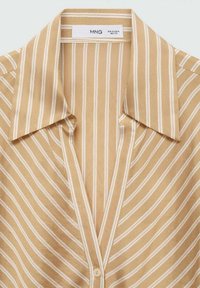Chemise beige à rayures verticales blanches, col large et pointu, avec un seul bouton visible sous le col.