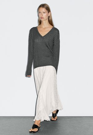 Femme portant un pull en V texturé gris foncé, une longue jupe blanche à motifs et des sandales noires à bout ouvert, se tenant contre un mur uni.